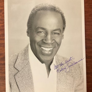 May include: Black and white photo of a man smiling. He is wearing a patterned jacket. The photo is signed "All the best, Robert Guillaume".