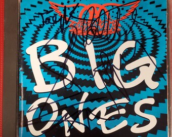 May include: A CD case with a blue and black background featuring the text "BIG ONES" in white. The band "Aerosmith" is printed in red on the case. The case is signed by the band members.