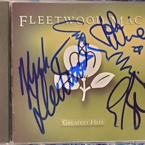 May include: A CD case with a green background featuring a white flower design. The text "FLEETWOOD MAC" is printed in blue at the top of the case. The text "GREATEST HITS" is printed in white at the bottom of the case. The case is signed in blue ink by Mick Fleetwood, Lindsey Buckingham, and Stevie Nicks.