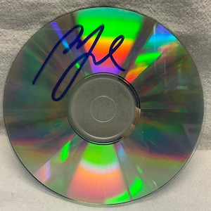 Könnte beinhalten: Eine silberne CD mit einem regenbogenfarbenen, irisierenden Schimmer. Die CD ist mit blauer Tinte mit dem Namen "Elle" signiert.