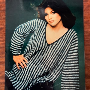 May include: A black and white striped sweater with a v-neck. The sweater is worn by a woman with dark hair and a neutral expression. The background is a dark green.