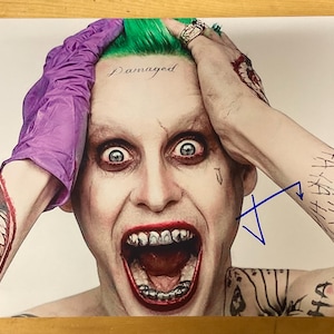 May include: A close-up of the Joker, a character from the Batman comics, with green hair and a purple glove. He is wearing a white shirt with a black and white pattern. The text "Damaged" is written on his forehead. The image is signed with a blue signature.