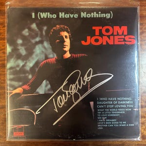 Può includere: Una copertina di un album in vinile in bianco e nero con Tom Jones. La copertina dell'album presenta una foto di Tom Jones con un maglione rosso. Il titolo dell'album è "I (Who Have Nothing)". La copertina dell'album è firmata con inchiostro argentato da Tom Jones.