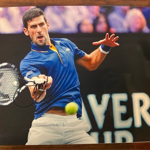 May include: A signed photograph of a tennis player wearing a blue shirt and white shorts, swinging a tennis racket. The player is on a tennis court with a crowd in the background. The photo is signed "Novak" in black ink.