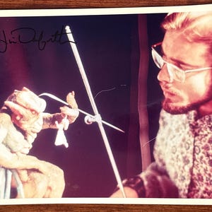 Puede incluir: Fotografía vintage que presenta a una persona con gafas y un títere. El títere es una criatura con un rostro y un disfraz detallados. La persona sostiene un dispositivo de control. La foto tiene una firma en la esquina superior izquierda.