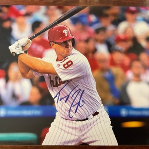 Può includere: Un giocatore di baseball con una maglia a righe bianche e rosse con il numero 8 sul davanti, oscilla una mazza. Il giocatore indossa un berretto da baseball rosso con una "P" bianca sul davanti. La firma del giocatore è visibile in inchiostro blu sulla maglia. Lo sfondo è un'immagine sfocata di una folla di persone.