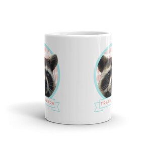 May include: White ceramic coffee mug with a raccoon face on each side. The raccoon on the left side has the text "Trash Panda" in a blue banner below it. The raccoon on the right side has the text "Trash Panda" in a blue banner below it.
