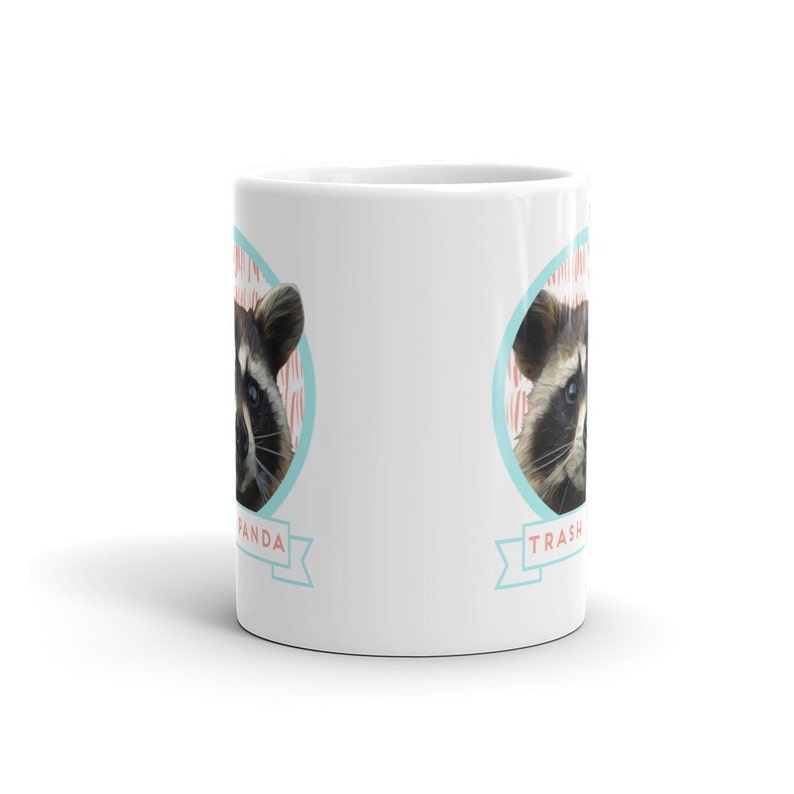 May include: White ceramic coffee mug with a raccoon face on each side. The raccoon on the left side has the text "Trash Panda" in a blue banner below it. The raccoon on the right side has the text "Trash Panda" in a blue banner below it.