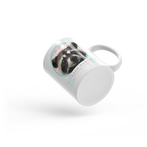 May include: White ceramic mug with a graphic of a raccoon's face and the text "Trash Panda" in a blue and pink banner.