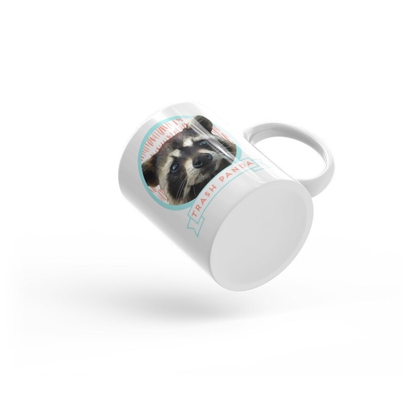 May include: White ceramic mug with a graphic of a raccoon's face and the text "Trash Panda" in a blue and pink banner.