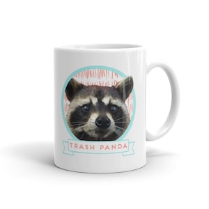 May include: White ceramic mug with a cartoon raccoon face and the text "Trash Panda" in a blue and white banner.