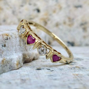 May include: A gold ring featuring a series of heart-shaped ruby and diamond accents. The ring's band is a simple, polished gold, and the gemstones are set in a repeating pattern. The ring is displayed on a textured, light-colored surface.