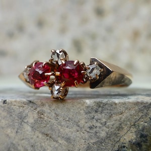 May include: A gold ring featuring three oval-cut, vibrant red gemstones, flanked by smaller, clear accent stones. The ring's design includes a geometric element on the sides, creating a unique and elegant look. The ring is set on a textured, gray surface.