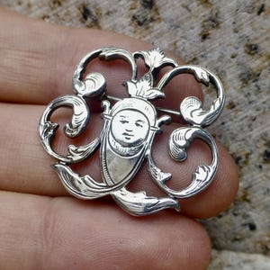 May include: A silver brooch with an elaborate design. The brooch showcases a central face with a crown, encircled by swirling, floral-inspired details. The pin is likely made of metal and has a classic, vintage appearance.