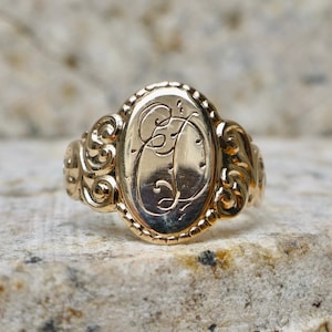 May include: A gold-toned ring featuring an oval-shaped face with a decorative beaded border. The centre of the oval is engraved with a stylised cursive letter D. The shoulders of the ring display intricate scrollwork patterns on both sides.