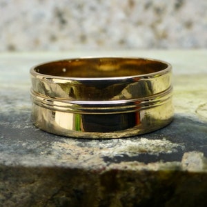 May include: A gold-colored ring with a wide band and two raised lines around the center. The ring is sitting on a textured surface.