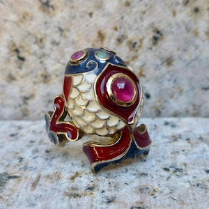 May include: An ornate ring in the shape of a fish, featuring a white body with red, blue, and gold detailing. The ring is adorned with small gemstones, including a central ruby. The design is intricate and eye-catching.