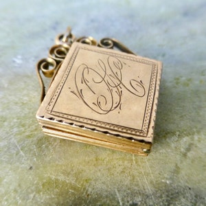 K&ouml;nnte beinhalten: Ein goldenes Medaillon mit einem zarten, verzierten Design. Das Medaillon hat einen Klappdeckel und eine kleine Schlaufe zum Anbringen einer Kette. Die Vorderseite des Medaillons ist mit den Initialen "C.L." graviert.