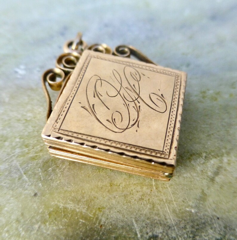 K&ouml;nnte beinhalten: Ein goldenes Medaillon mit einem zarten, verzierten Design. Das Medaillon hat einen Klappdeckel und eine kleine Schlaufe zum Anbringen einer Kette. Die Vorderseite des Medaillons ist mit den Initialen "C.L." graviert.