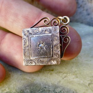 K&ouml;nnte beinhalten: Ein goldenes Medaillon mit einem dekorativen Filigranmuster und einem einzelnen diamantf&ouml;rmigen Stein in der Mitte. Das Medaillon ist geschlossen und hat eine quadratische Form.