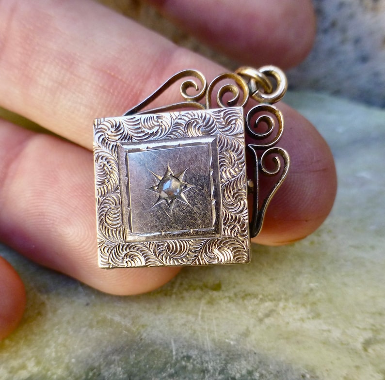 K&ouml;nnte beinhalten: Ein goldenes Medaillon mit einem dekorativen Filigranmuster und einem einzelnen diamantf&ouml;rmigen Stein in der Mitte. Das Medaillon ist geschlossen und hat eine quadratische Form.