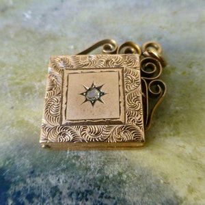 K&ouml;nnte beinhalten: Ein goldenes Medaillon mit einem diamantf&ouml;rmigen Stein in der Mitte. Das Medaillon hat ein dekoratives, graviertes Design und eine filigranartige &Ouml;se.