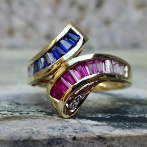 May include: A bypass ring crafted from yellow metal featuring two overlapping bands. One band is channel-set with rectangular baguette-cut blue gemstones, while the other band is channel-set with rectangular baguette-cut pink and red gemstones.