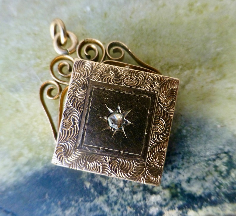 K&ouml;nnte beinhalten: Ein goldenes Medaillon mit einem dekorativen Filigranmuster und einem einzigen diamantf&ouml;rmigen Stein in der Mitte.