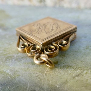 K&ouml;nnte beinhalten: Ein goldener Anh&auml;nger mit quadratischer Form und einem komplizierten Schn&ouml;rkeldesign. Der Anh&auml;nger hat einen Klappdeckel und eine Schlaufe zum Anbringen einer Kette. Der Anh&auml;nger ist mit einem Monogramm graviert.