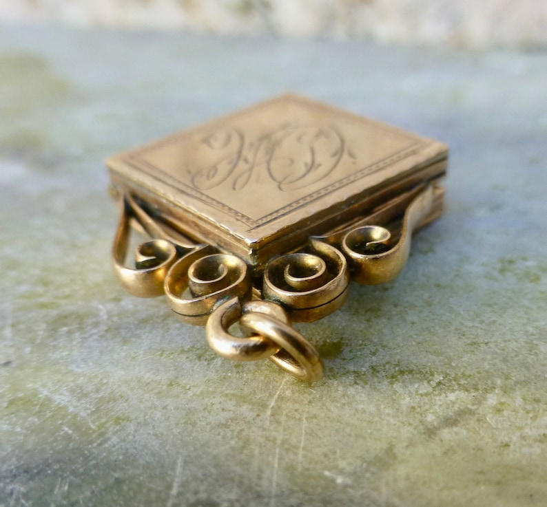 K&ouml;nnte beinhalten: Ein goldener Anh&auml;nger mit quadratischer Form und einem komplizierten Schn&ouml;rkeldesign. Der Anh&auml;nger hat einen Klappdeckel und eine Schlaufe zum Anbringen einer Kette. Der Anh&auml;nger ist mit einem Monogramm graviert.