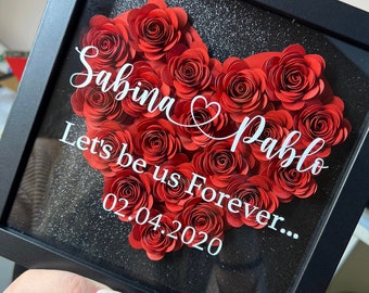 May include: A black framed shadow box with a red paper flower heart. The heart is made of red paper roses and has the text "Sabina & Pablo Let's be us Forever... 02.04.2020" written in white.