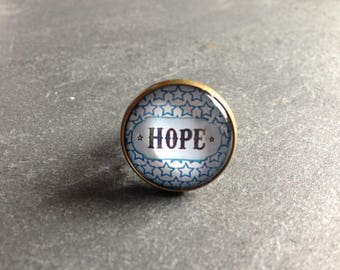 Puede incluir: Un cabujón redondo de vidrio azul y blanco con un engaste de metal dorado. El cabujón presenta un patrón de estrellas blancas y la palabra "HOPE" en letras negras.