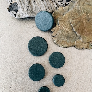 Op de afbeelding: Zes donkergroene ronde schijven van verschillende grootte op een lichtbeige, getextureerd oppervlak.