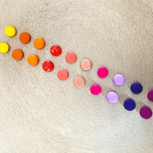 Könnte beinhalten: Ein Regenbogen-Farbverlauf aus runden, bunten Knöpfen, die in einer geschwungenen Linie auf einer hellbraunen, strukturierten Oberfläche angeordnet sind.