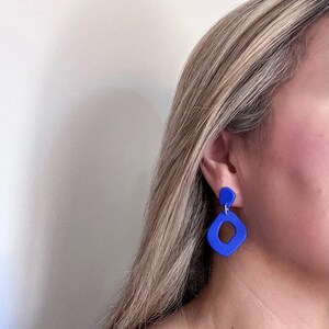 May include: A pair of blue geometric earrings with a square shape and a smaller circle shape. The earrings are made of a smooth, matte material.