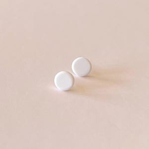 Op de afbeelding: Twee kleine, witte, ronde oorstekers tegen een zachtroze achtergrond. De oorbellen hebben een gladde, matte afwerking en zijn gelijkmatig verdeeld. Het minimalistische ontwerp suggereert een eenvoudige, elegante stijl.