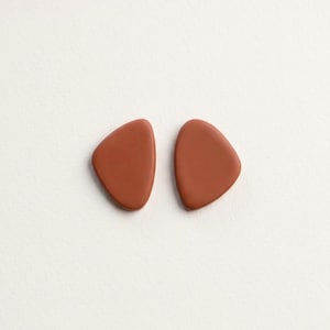 Peut inclure: Une paire de boucles d'oreilles à tige présentant des surfaces lisses et mates de couleur terre cuite. Chaque boucle d'oreille a une forme triangulaire arrondie asymétrique distincte avec des bords doux et incurvés.