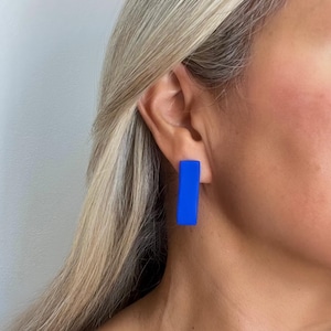 May include: A pair of vibrant blue rectangular earrings. The earrings are a solid, bold blue color and are worn on the ear. The image shows a close-up of the earrings against a neutral background.