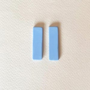Puede incluir: Dos pendientes rectangulares de color azul claro. Cada pendiente es de color azul pastel liso con un acabado liso y mate. Los pendientes están colocados verticalmente, uno al lado del otro, sobre un fondo blanco.