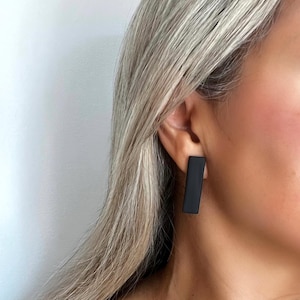Peut inclure: Gros plan d'une boucle d'oreille rectangulaire noire. La boucle d'oreille est de couleur noire mate unie et présente un design simple et minimaliste. La boucle d'oreille est portée à l'oreille d'une personne aux cheveux blonds.