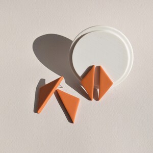 May include: A pair of orange triangle shaped earrings. The earrings are made of polymer clay and have a matte finish.