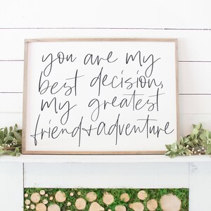 May include: A white wooden sign with black calligraphy that reads "You are my best decision, my greatest friend + adventure". The sign is sitting on a white mantel with green foliage on either side.