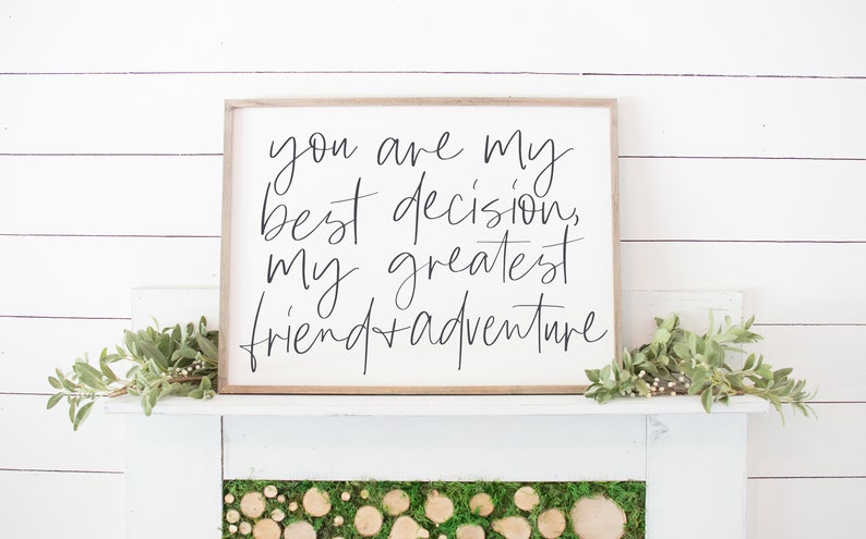May include: A white wooden sign with black calligraphy that reads "You are my best decision, my greatest friend + adventure". The sign is sitting on a white mantel with green foliage on either side.