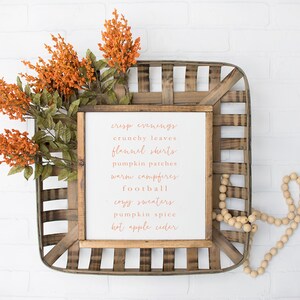 May include: A white wooden sign with the text "crisp evenings, crunchy leaves, flannel shirts, pumpkin patches, warm campfires, football, cozy sweaters, pumpkin spice, hot apple cider" in orange lettering. The sign is in a wooden basket with orange flowers and wooden beads.