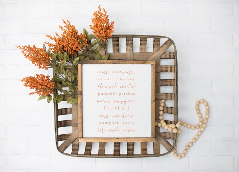 May include: A white wooden sign with the text "crisp evenings, crunchy leaves, flannel shirts, pumpkin patches, warm campfires, football, cozy sweaters, pumpkin spice, hot apple cider" in orange lettering. The sign is in a wooden basket with orange flowers and wooden beads.