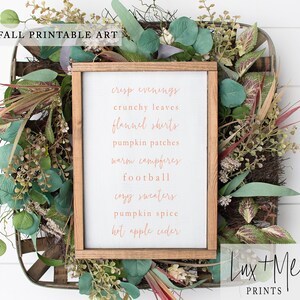 May include: A white wood sign with a brown frame, featuring a list of fall-themed words in a copper-colored font. The words are: crisp evenings, crunchy leaves, flannel shirts, pumpkin patches, warm campfires, football, cozy sweaters, pumpkin spice, hot apple cider. The sign is surrounded by greenery.
