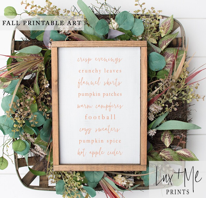May include: A white wood sign with a brown frame, featuring a list of fall-themed words in a copper-colored font. The words are: crisp evenings, crunchy leaves, flannel shirts, pumpkin patches, warm campfires, football, cozy sweaters, pumpkin spice, hot apple cider. The sign is surrounded by greenery.