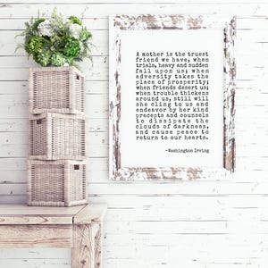 May include: A white framed print with a black and white text quote about a mother being the truest friend. The quote is by Washington Irving. The print is hanging on a white wood wall with a wicker basket in the foreground.