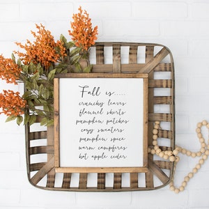 May include: A framed sign with the text "Fall is... crunchy leaves, flannel shirts, pumpkin patches, cozy sweaters, pumpkin spice, warm campfires, hot apple cider" on a white background. The sign is sitting in a wooden basket with orange flowers and wooden beads.