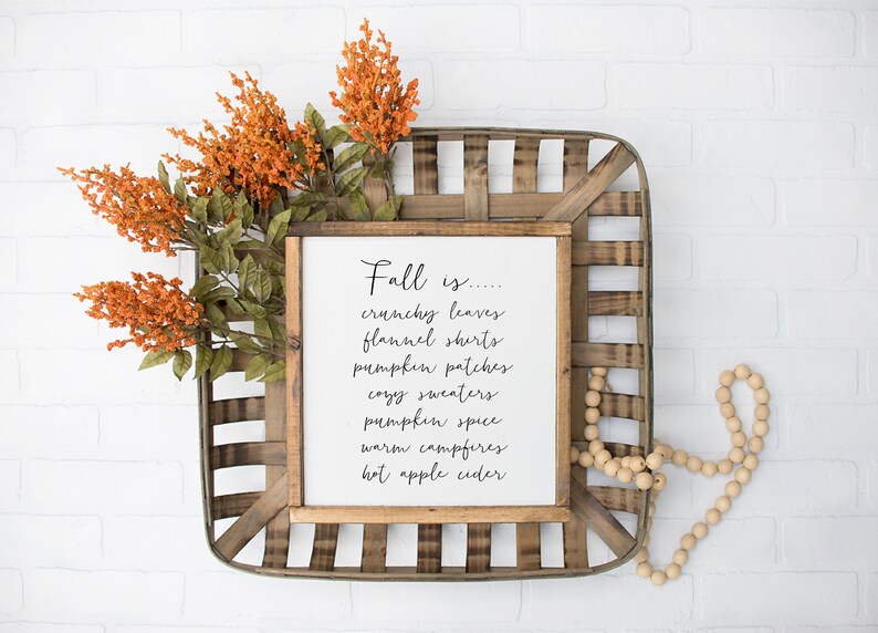 May include: A framed sign with the text "Fall is... crunchy leaves, flannel shirts, pumpkin patches, cozy sweaters, pumpkin spice, warm campfires, hot apple cider" on a white background. The sign is sitting in a wooden basket with orange flowers and wooden beads.